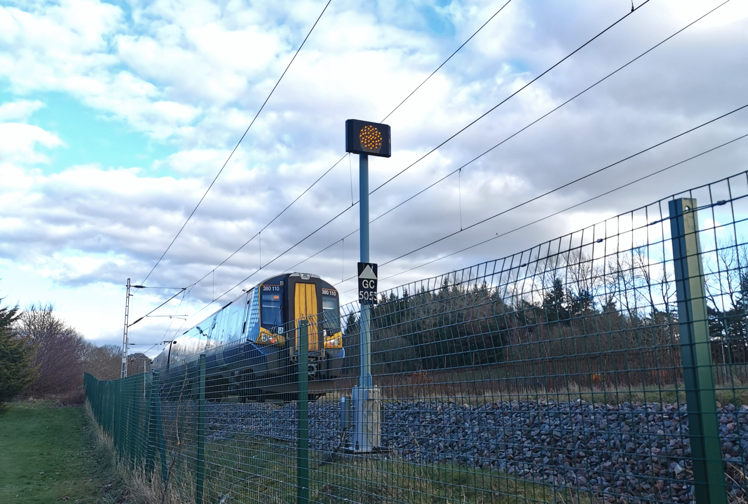 A ScotRail class 380 train viewed from bellow, behind a fence, passing an amber signal on a cloudy day