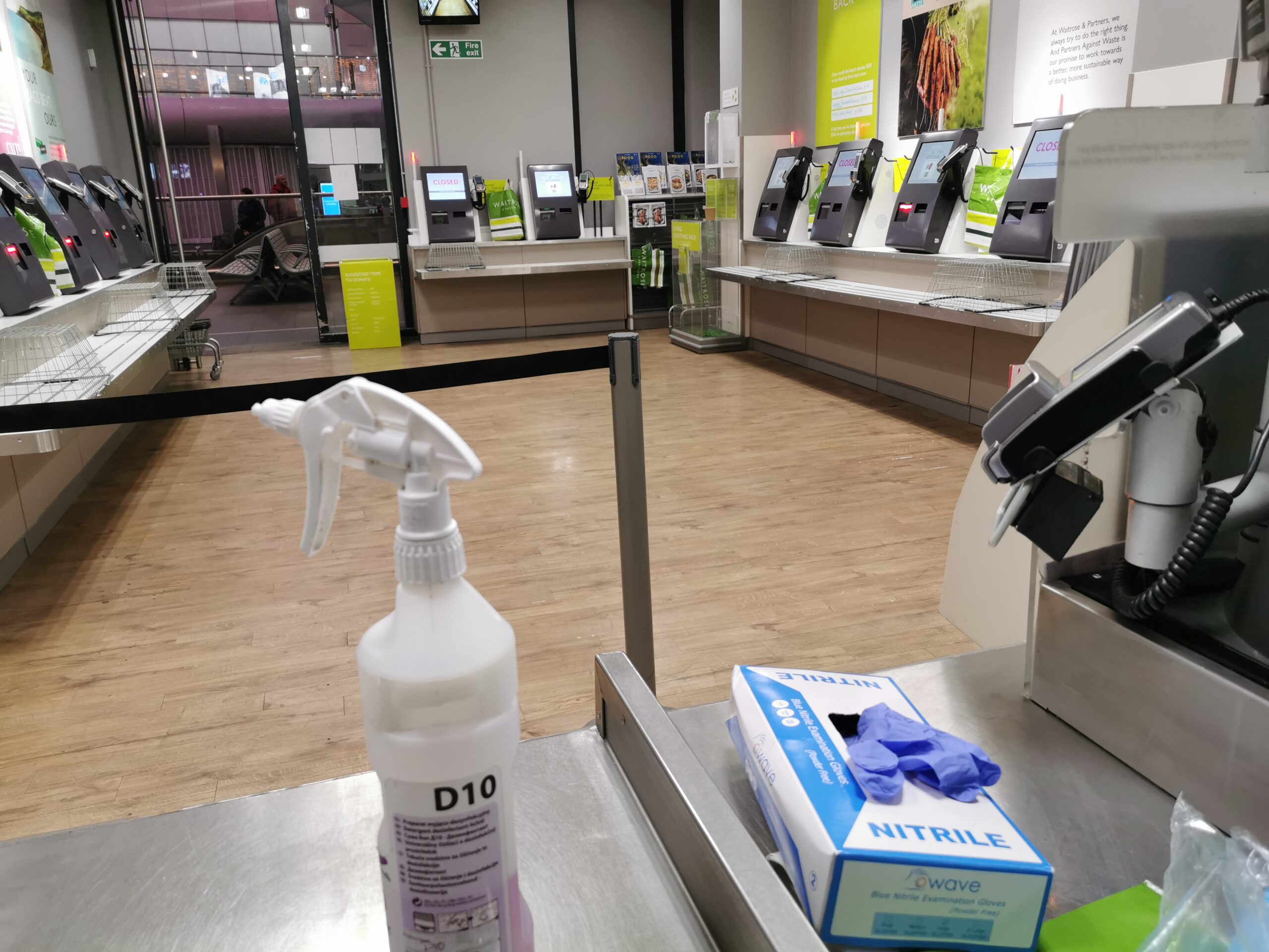 View from behind the tills in Waitrose Kings Cross, 2020 layout. A bank of small self checkouts are alternatingly disabled to enforce social distancing. a D10 disinfectant bottle and pack of gloves is in shot on the counter. The station is almost empty of passengers