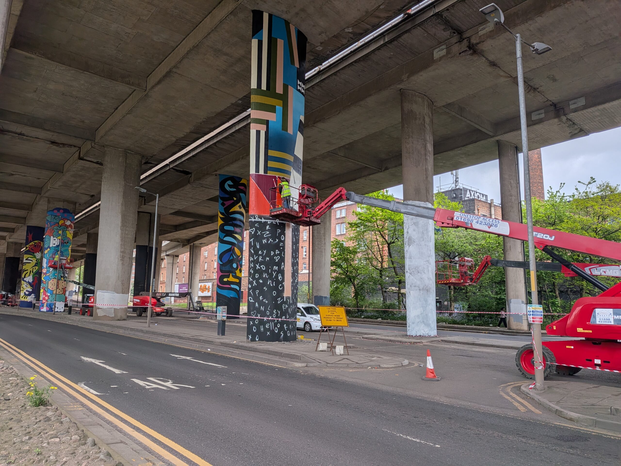 A pedestrian view under the Kingston bridge viaducts on the north side. The pillars are being painted with steat art, a crane lifts an aritst into place in the foreground. Trees are visible behind the structure and a lone weed poking through in the foreground. The roof of the structure is clearly distressed, a constrast with the art literally covering over the concrete bellow.